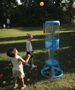 Two children play with a tall plastic game outdoors, tossing balls, on a grassy field with trees in the background.