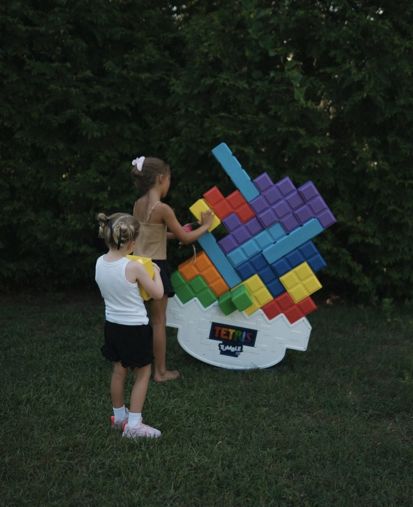Two girls playing Tetris outside with a large colorful plastic Tetris tower on grass near green bushes.