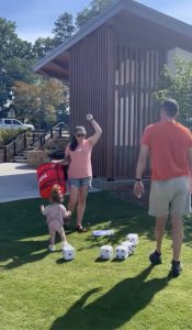 Three people play with oversized dice on grass near a building; the woman is about to throw a die.