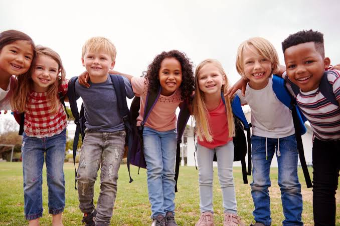 images Seven smiling children with backpacks stand together outside, arms around each other, looking at the camera.