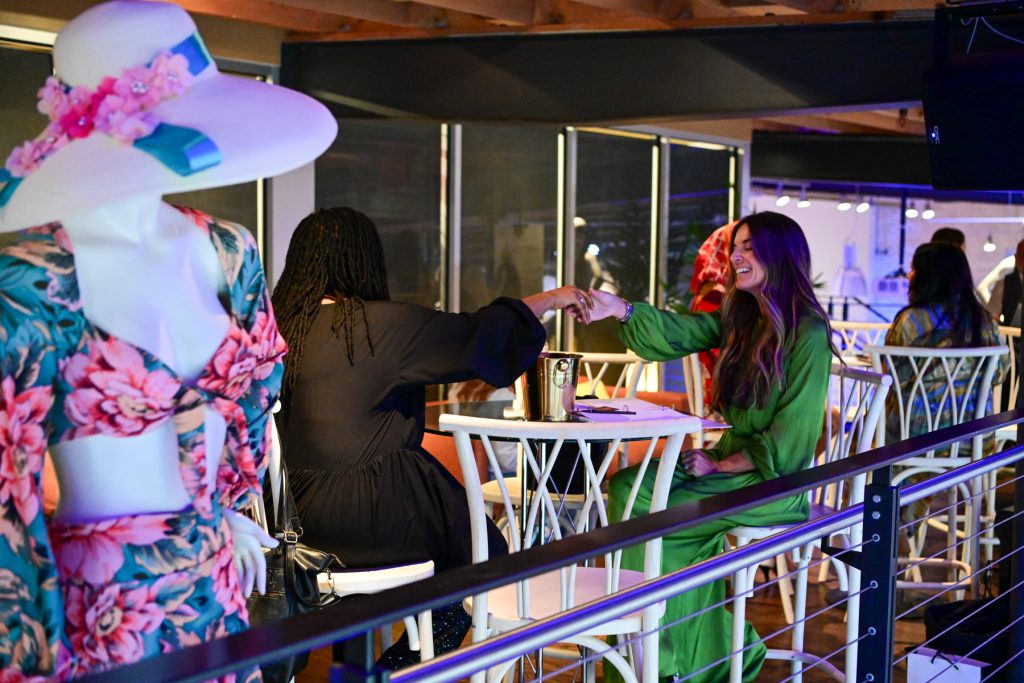 Two women fist bump at a cafe table; a floral-dressed mannequin in a hat is in the foreground.