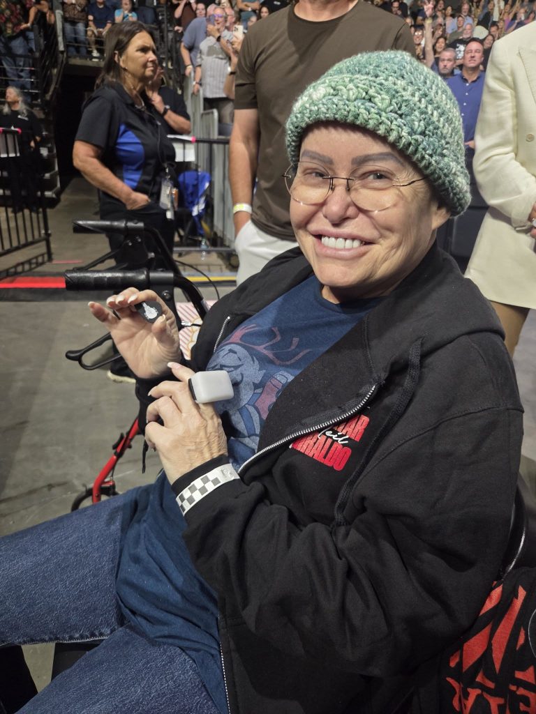 Smiling person in glasses and a green beanie sits in a crowd, holding a small device, wearing a black hoodie.