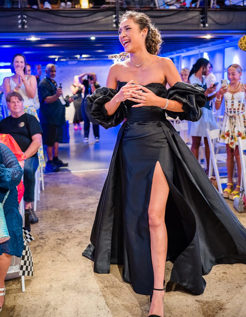 Smiling woman in a black dress with puffy sleeves walks down an aisle as people applaud in a bright room.