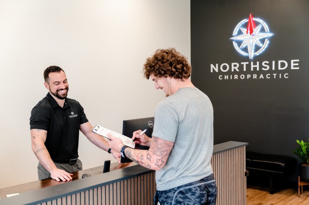 A man signs a form at a chiropractic clinic reception while the receptionist smiles behind the desk.