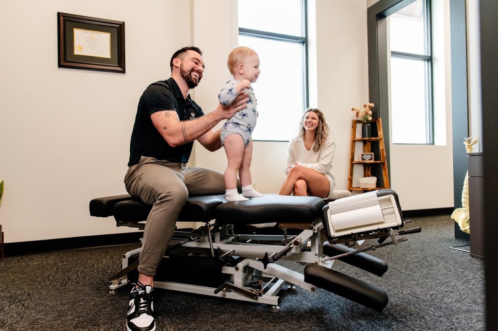 A man helps a baby stand on a chiropractic table while a woman smiles and watches nearby.