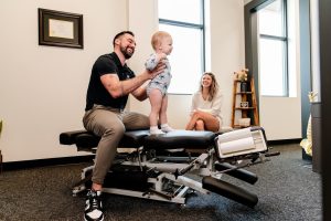 A man helps a baby stand on a chiropractic table while a woman smiles and watches nearby.