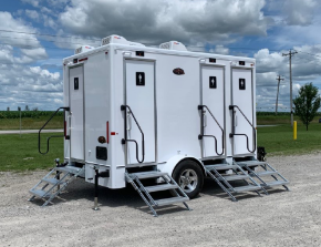 White portable restroom trailer with three doors and metal steps, parked on gravel under a cloudy sky.