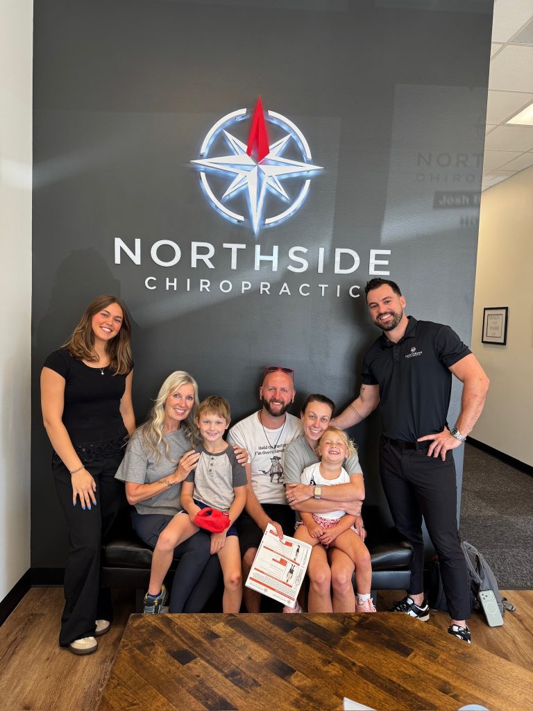 A smiling family and two staff pose in front of a Northside Chiropractic sign in an office lobby.