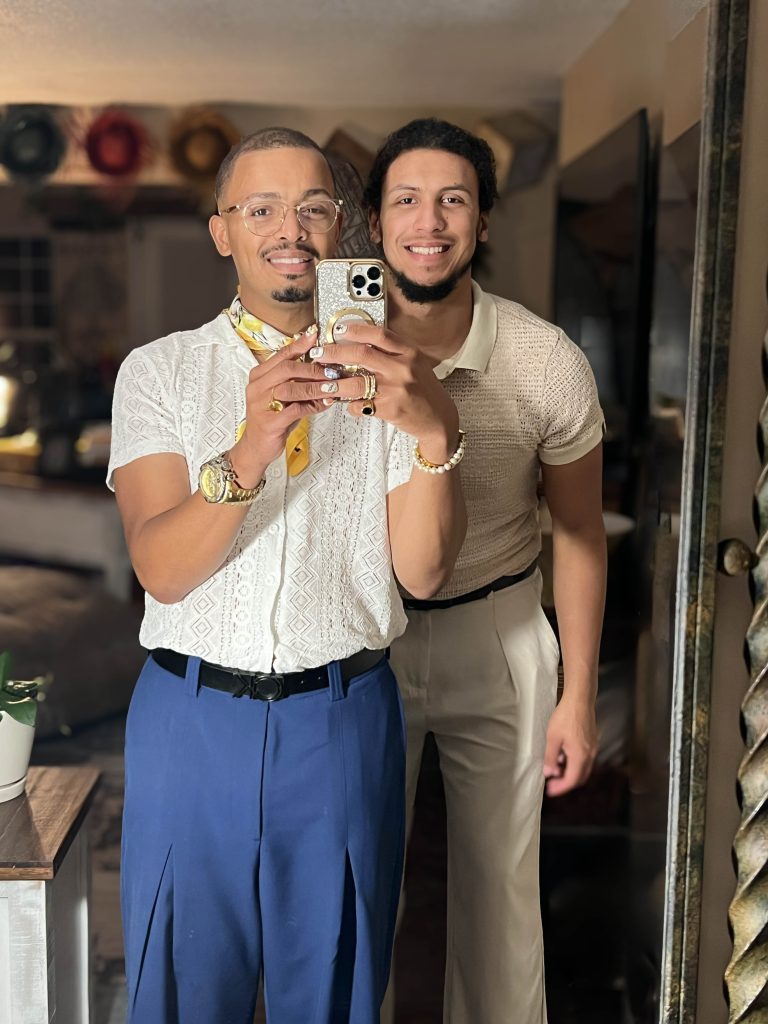 Two men smiling and posing for a mirror selfie in stylish outfits inside a warmly lit room.