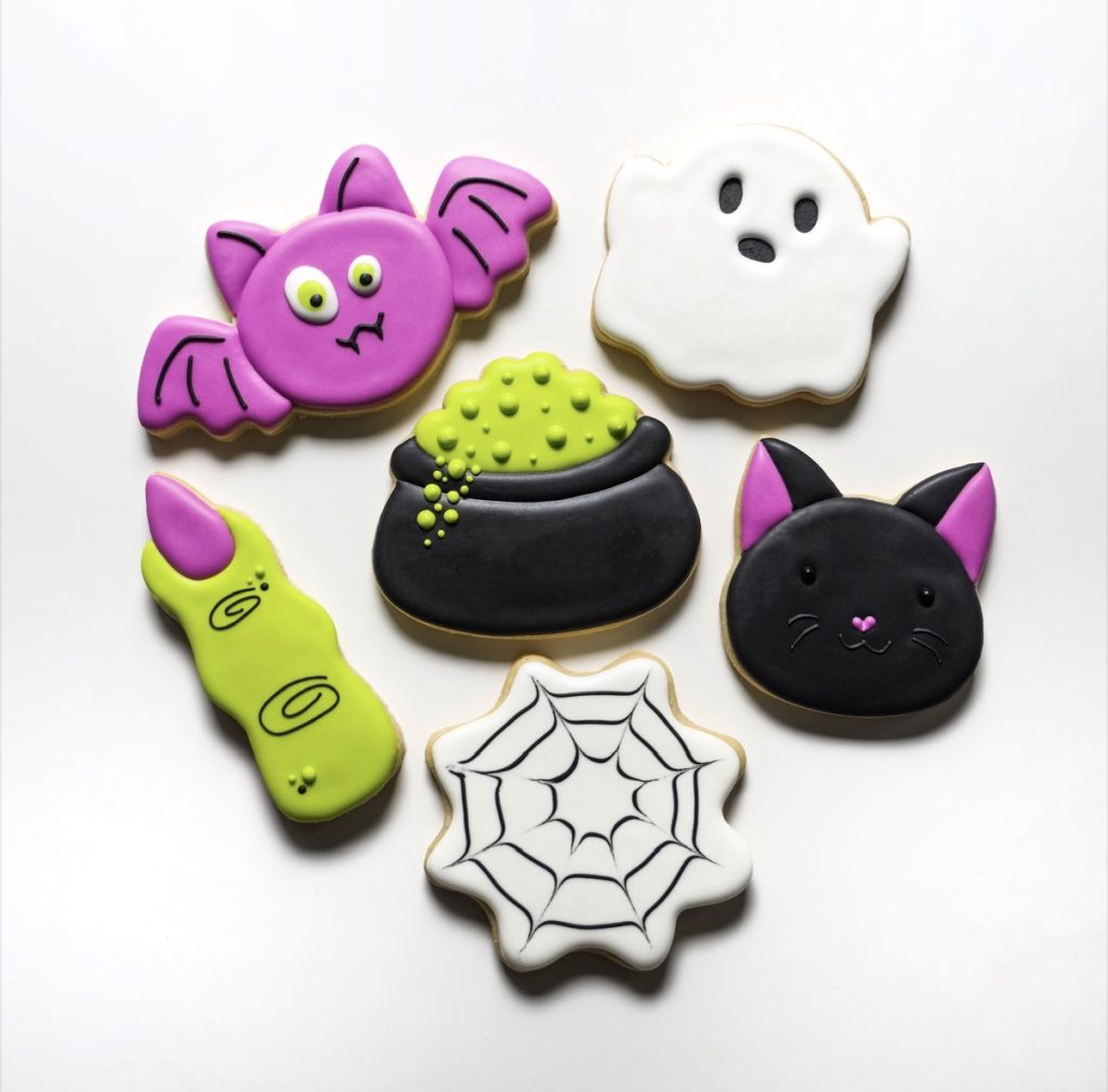 Six Halloween cookies: a bat, ghost, bubbling cauldron, cat, witch finger, and spider web on white background.