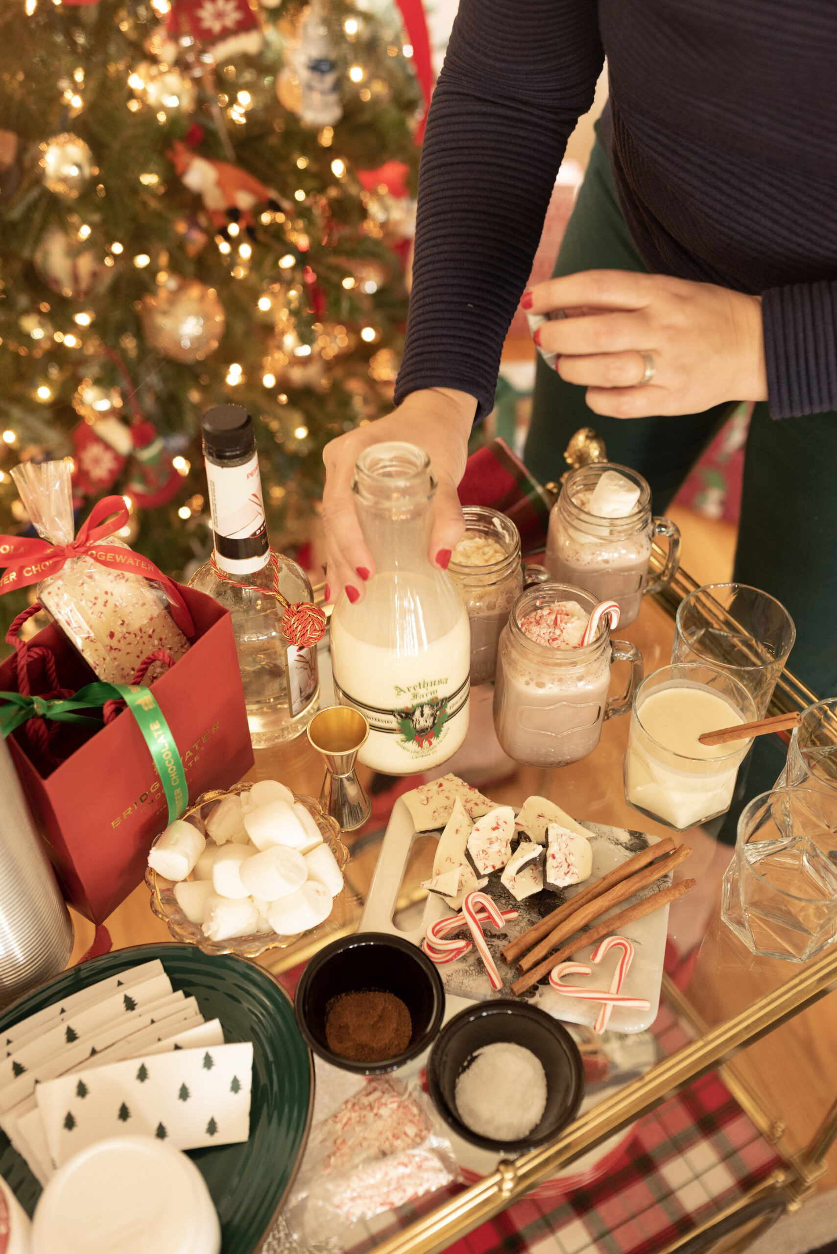 A person prepares holiday drinks with eggnog, marshmallows, candy canes, and treats near a decorated Christmas tree.