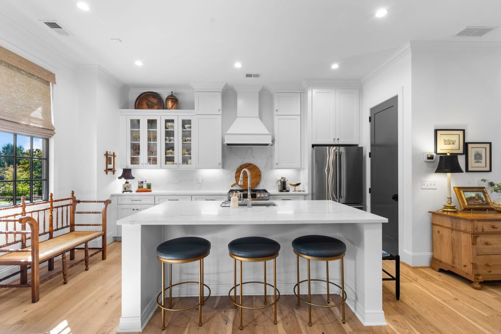 Modern white kitchen with a marble-topped island, three bar stools, and a wooden bench by the window.