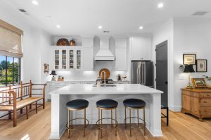 Modern white kitchen with a marble-topped island, three bar stools, and a wooden bench by the window.