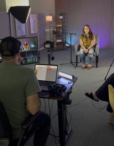 A person sits in a studio chair being filmed for an interview while a producer monitors the recording on a laptop.