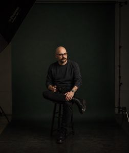 A bald man with a mustache, wearing glasses and a sweater, sitting on a stool and holding a camera.