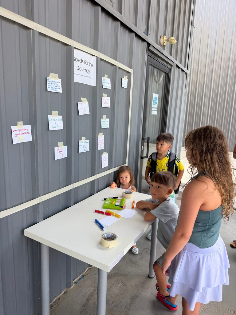 Children and an adult write notes at a white table under a board with cards taped to a metal wall.