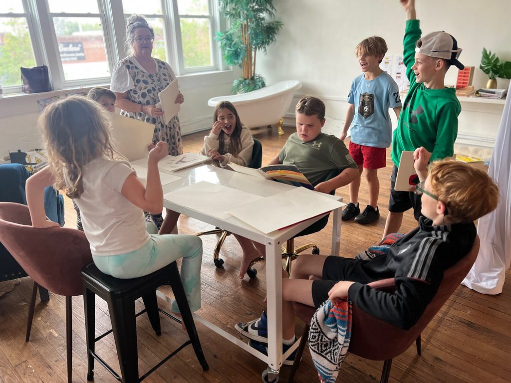 A group of children sits around a table, laughing and raising hands, while a woman stands holding papers.