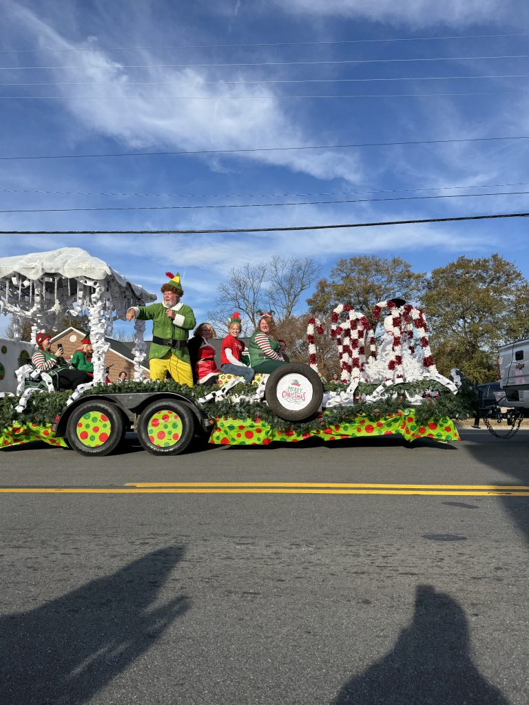 People in festive costumes ride a decorated Christmas parade float on a sunny day, with trees in the background.