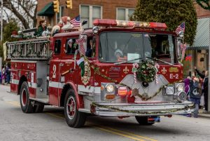 A decorated red fire truck in a parade, with people on top waving and festive wreaths on the front.