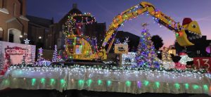 Excavator decorated with Christmas lights and ornaments, next to trees and North Pole sign in a parade float.
