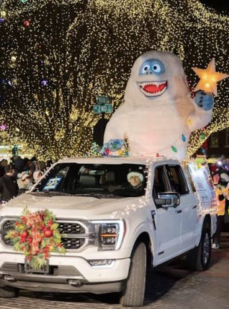 White truck with wreath, festive lights, and large smiling yeti holding a star on top during a parade.
