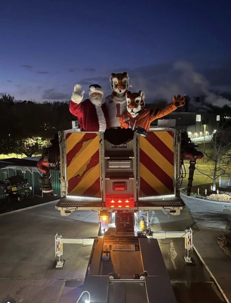 Santa and two tiger mascots stand on a fire truck ladder platform at night, waving.