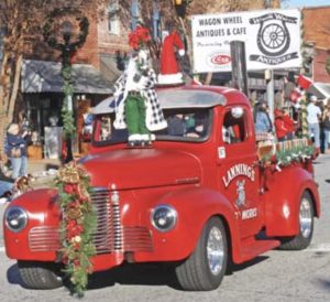 Red vintage truck decorated for Christmas drives in a parade, with festive ornaments and people watching nearby.