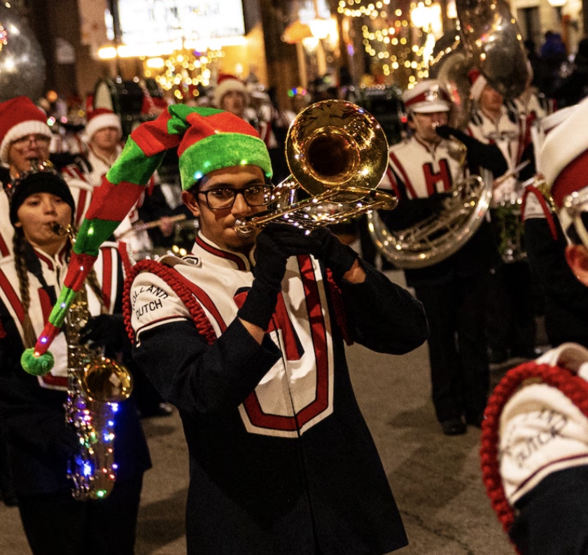 Marching band in uniform plays instruments at night, with one member wearing a festive elf hat.