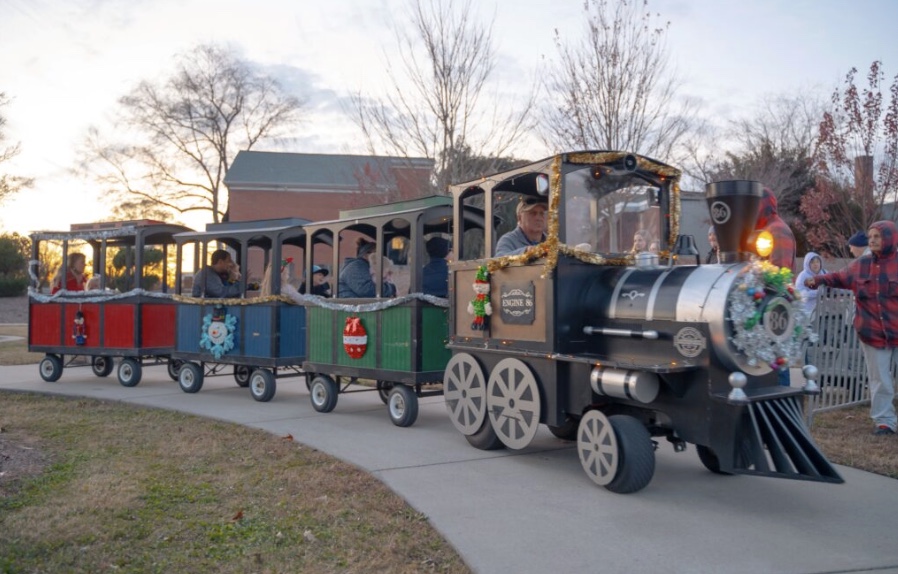 A small festive train with adults and children rides on a sidewalk, decorated for the holidays.