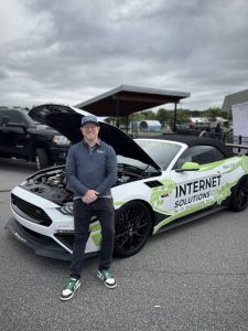 A man stands in front of a white Ford Mustang with the hood up, branded with “Internet Solutions Co” graphics.