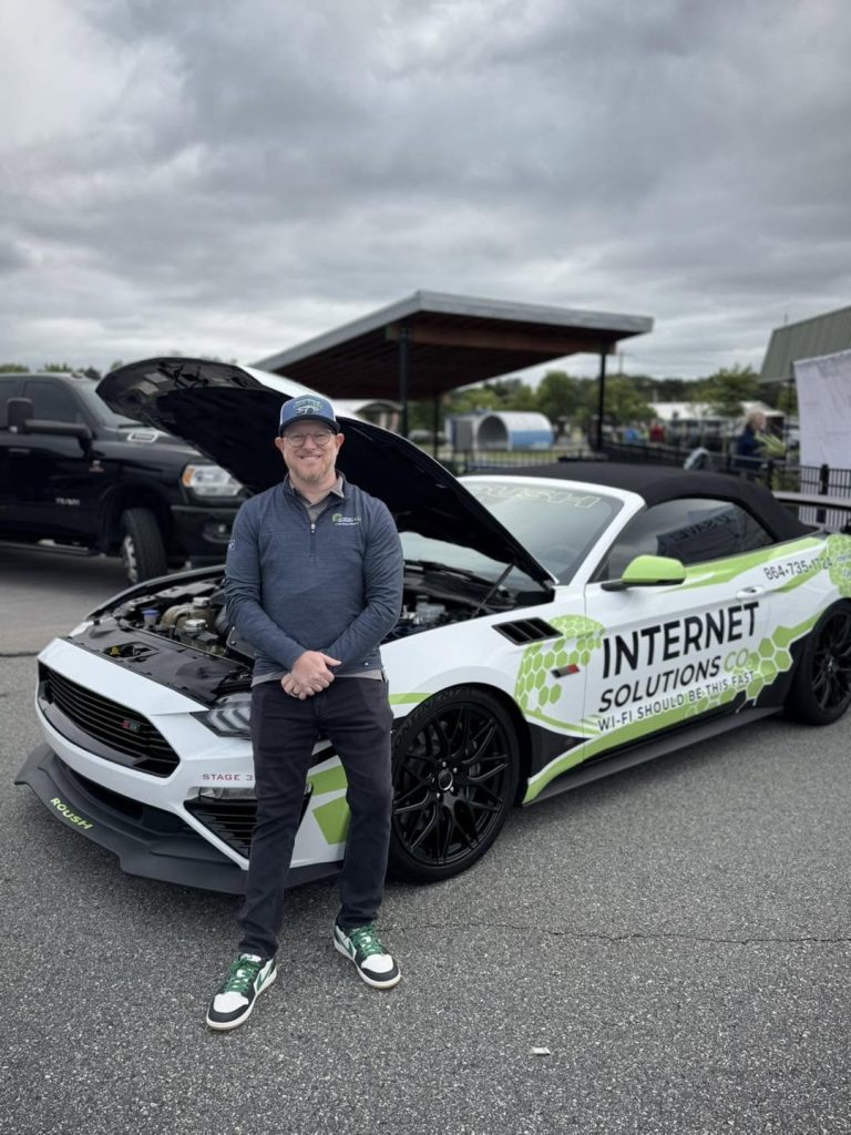 A man stands in front of a white Ford Mustang with the hood up, branded with “Internet Solutions Co” graphics.