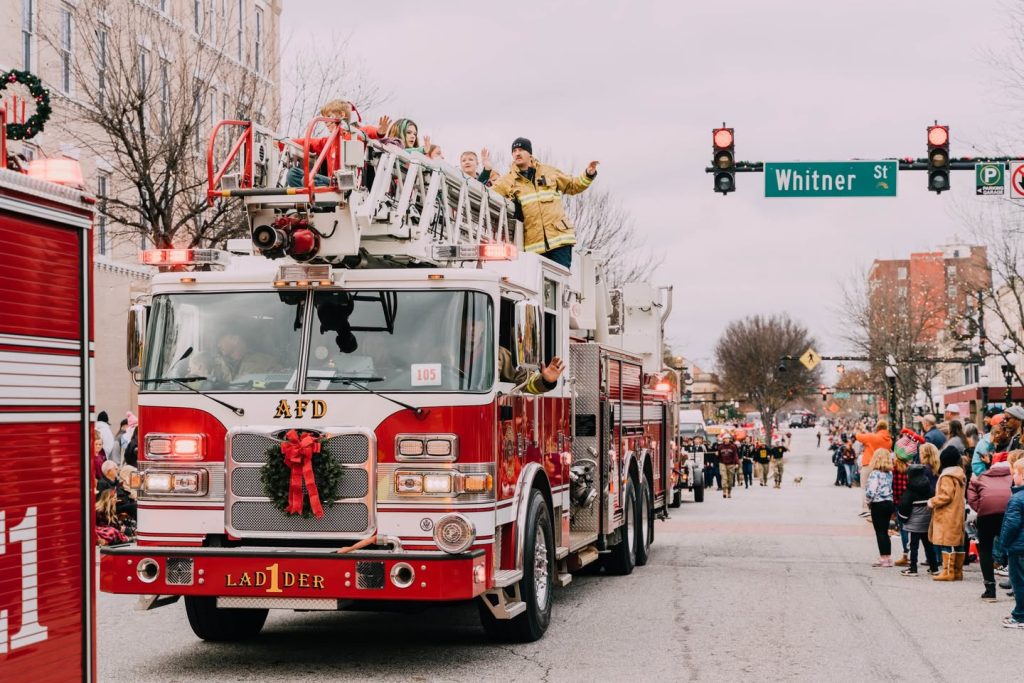 A fire truck decorated for Christmas leads a parade, with people waving on top and crowds lining the street.