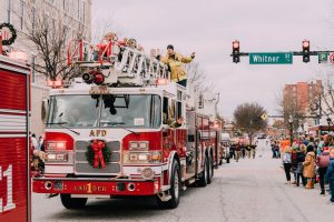A fire truck decorated for Christmas leads a parade, with people waving on top and crowds lining the street.